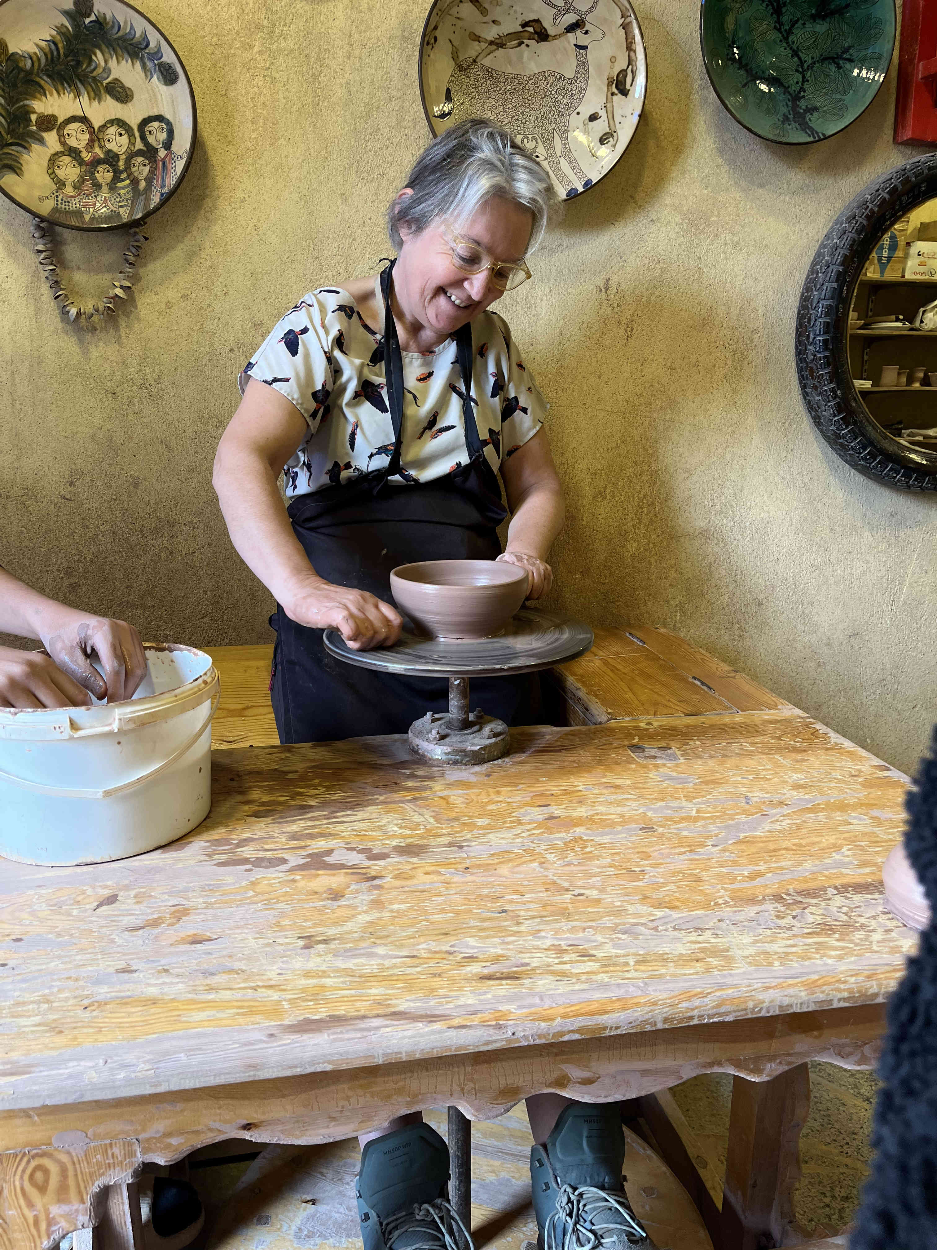 A smiling woman in a workshop demonstrates making pottery on a traditional wheel