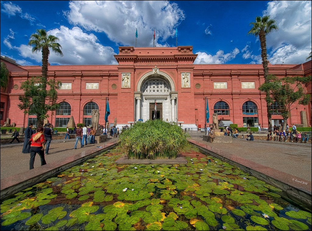 Extérieur du bâtiment historique en brique rouge du Musée Égyptien du Caire (Place Tahrir), avec un bassin de nénuphars et des palmiers au premier plan
