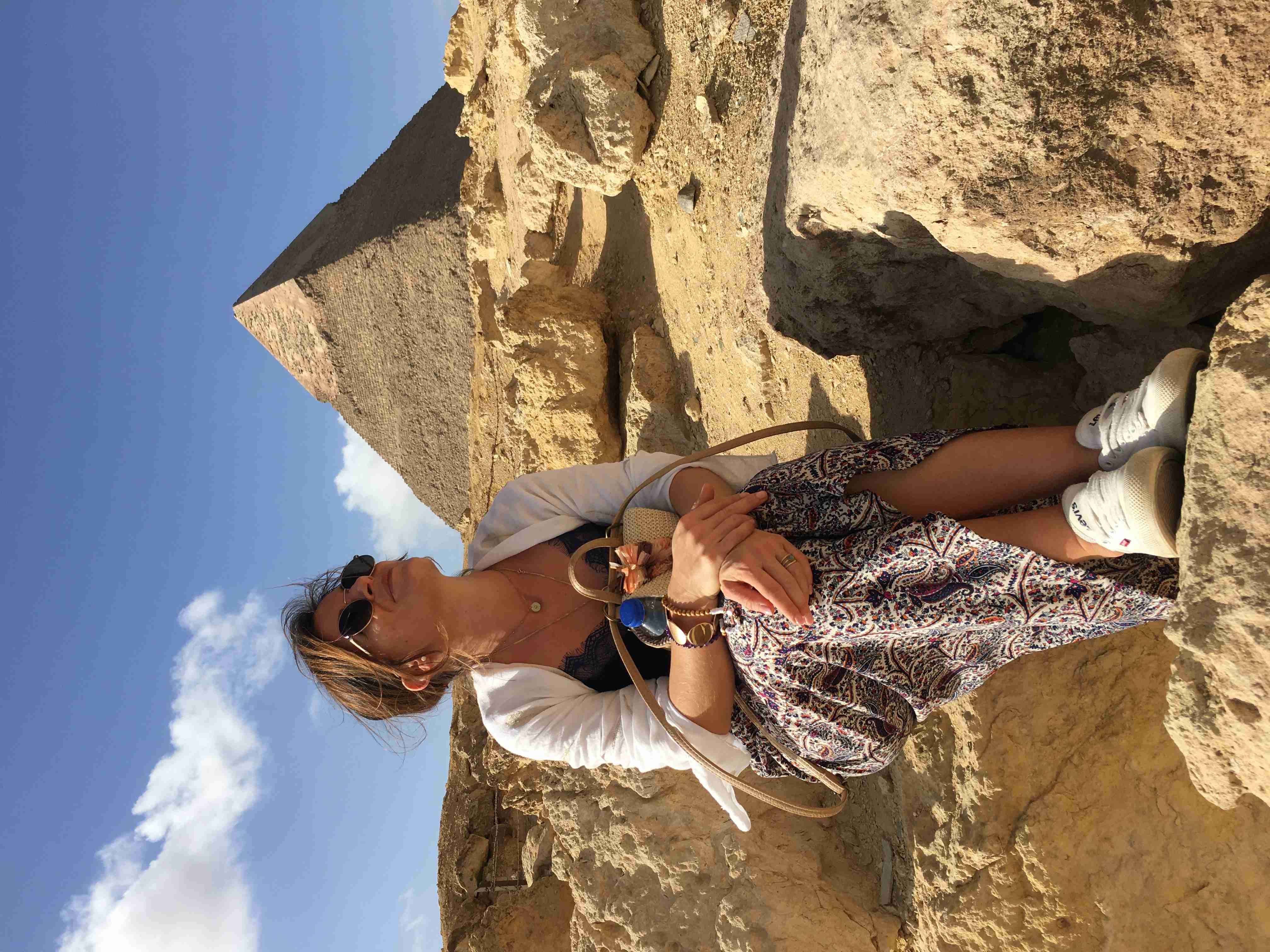 Une femme assise sur un sol rocailleux dans le désert égyptien, avec la pyramide bien conservée de Mykérinos (ou Khéphren) dans la nécropole de Gizeh visible derrière elle.