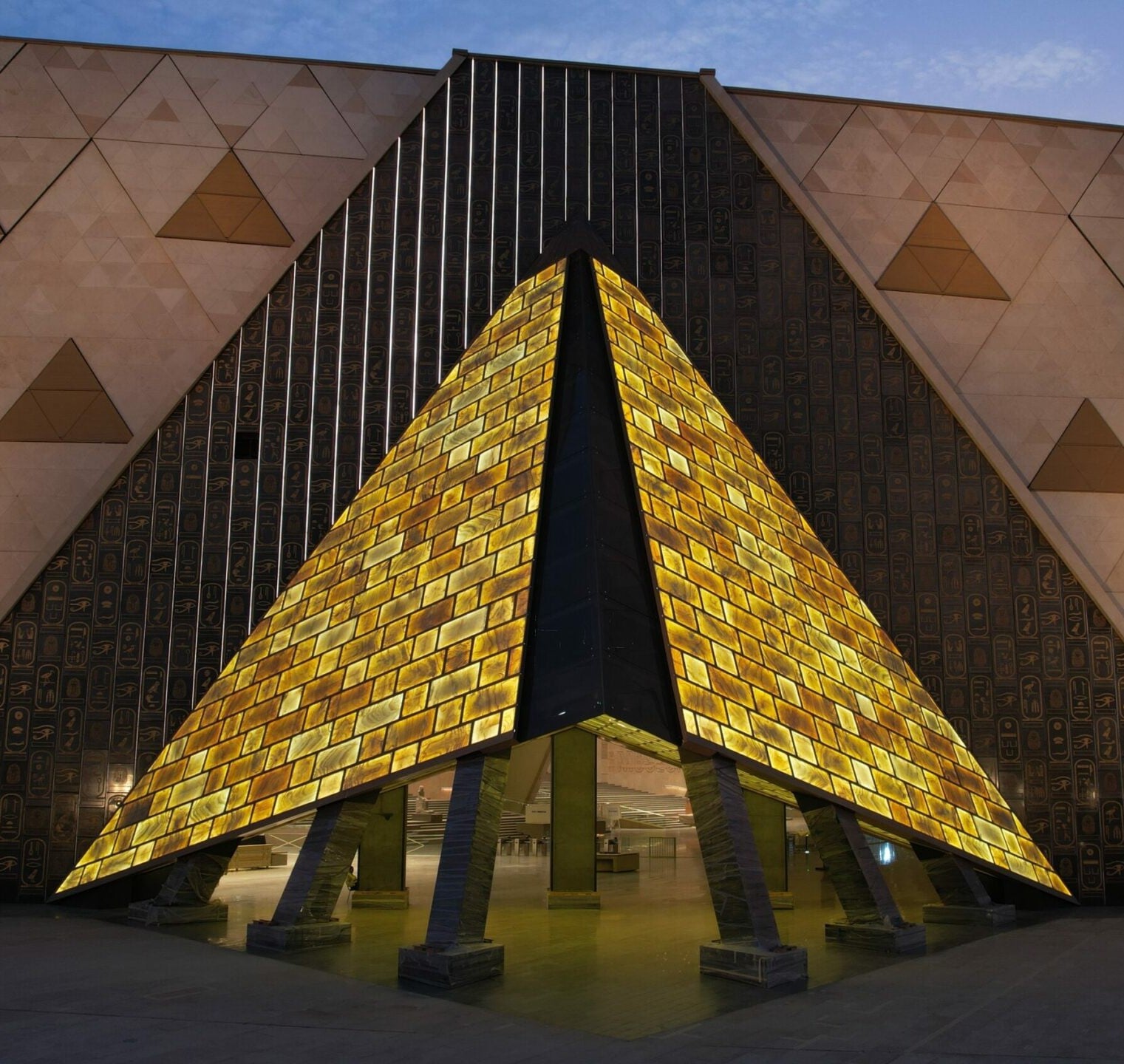 La façade et le porche d'entrée illuminés en forme de pyramide dorée du Grand Musée Égyptien (GEM) moderne, la nuit, présentent un mur orné de hiéroglyphes.