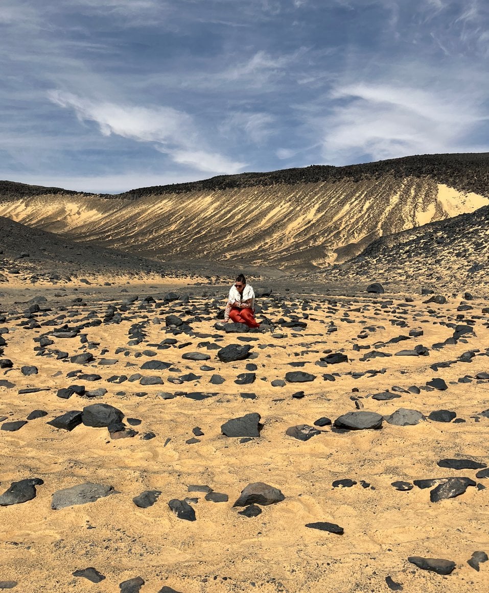 Une personne assise au milieu d'un cercle de pierres volcaniques noires sur le sable jaune-orangé du Désert Noir en Égypte, avec des pentes sombres et érodées en arrière-plan