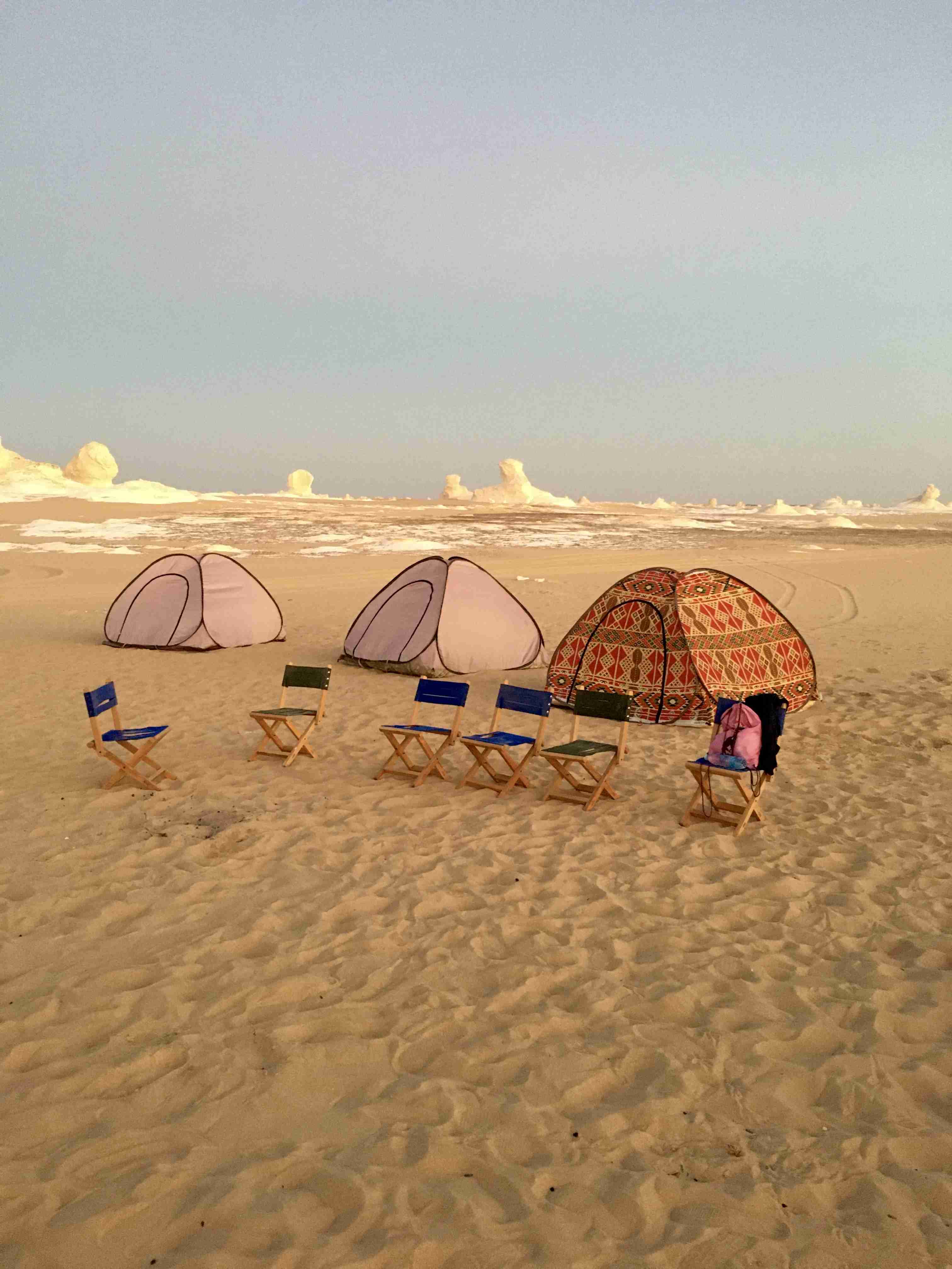 Un camping pittoresque au crépuscule dans le Désert Blanc, avec trois tentes et plusieurs petites chaises pliantes disposées sur le sable blanc-crème, avec des formations calcaires lointaines