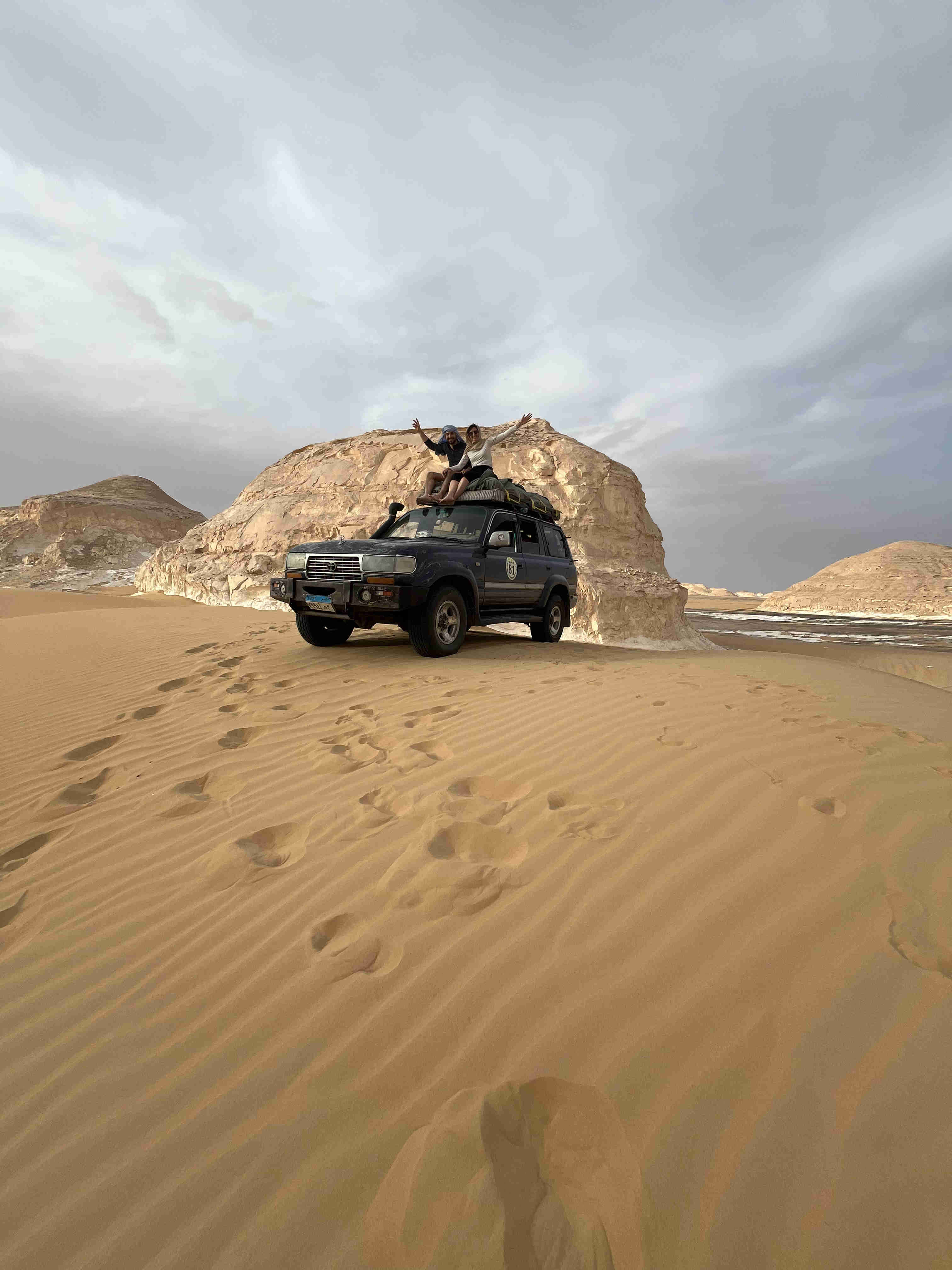 A 4x4 vehicle with travelers on the roof is parked atop a golden sand dune, surrounded by the towering, white chalk formations of the White Desert National Park