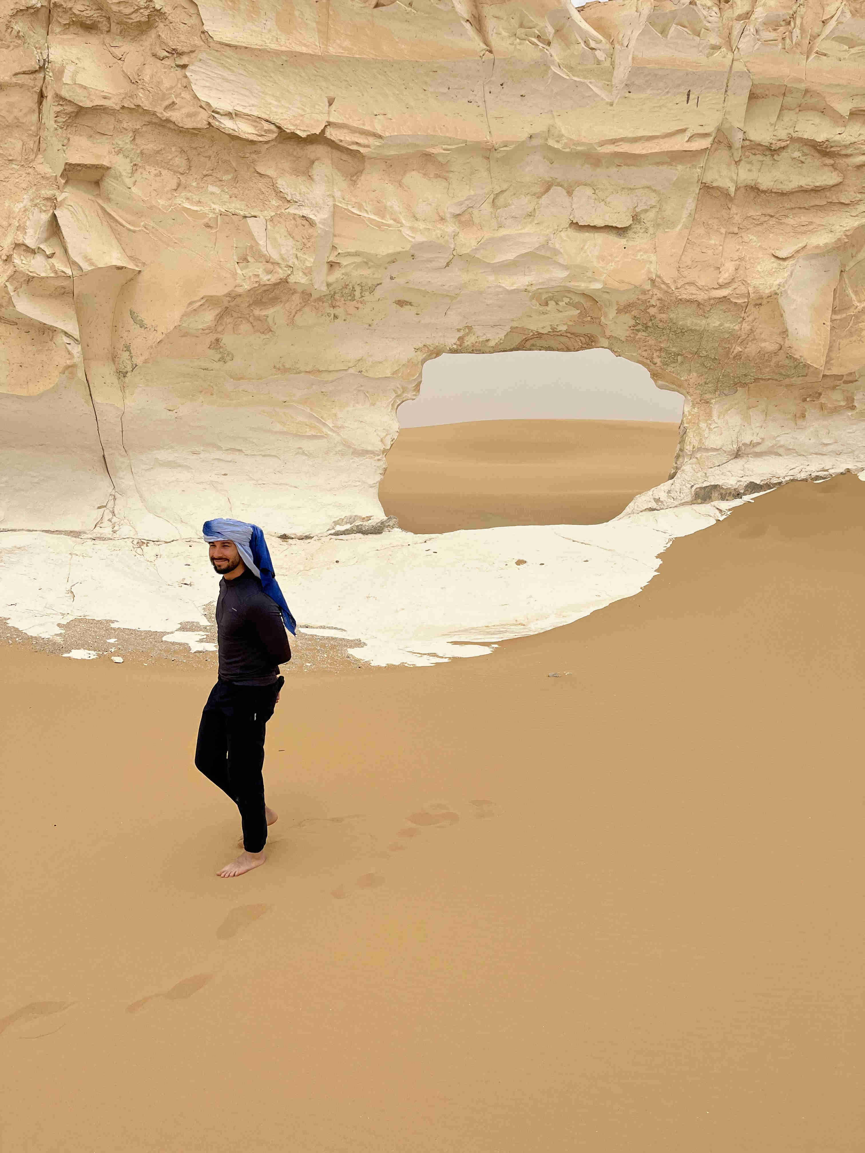 A man walking barefoot on golden sand beneath a naturally eroded white chalk archway in the White Desert, with the distant desert visible through the opening