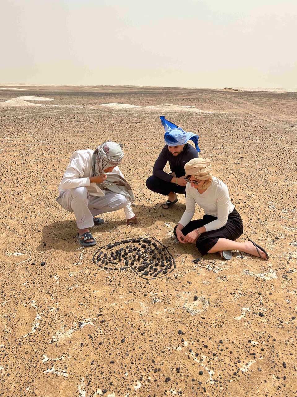 Two tourists and a local guide kneeling in the Black Desert of Egypt, examining a heart shape made from dark volcanic stones on the light brown desert floor