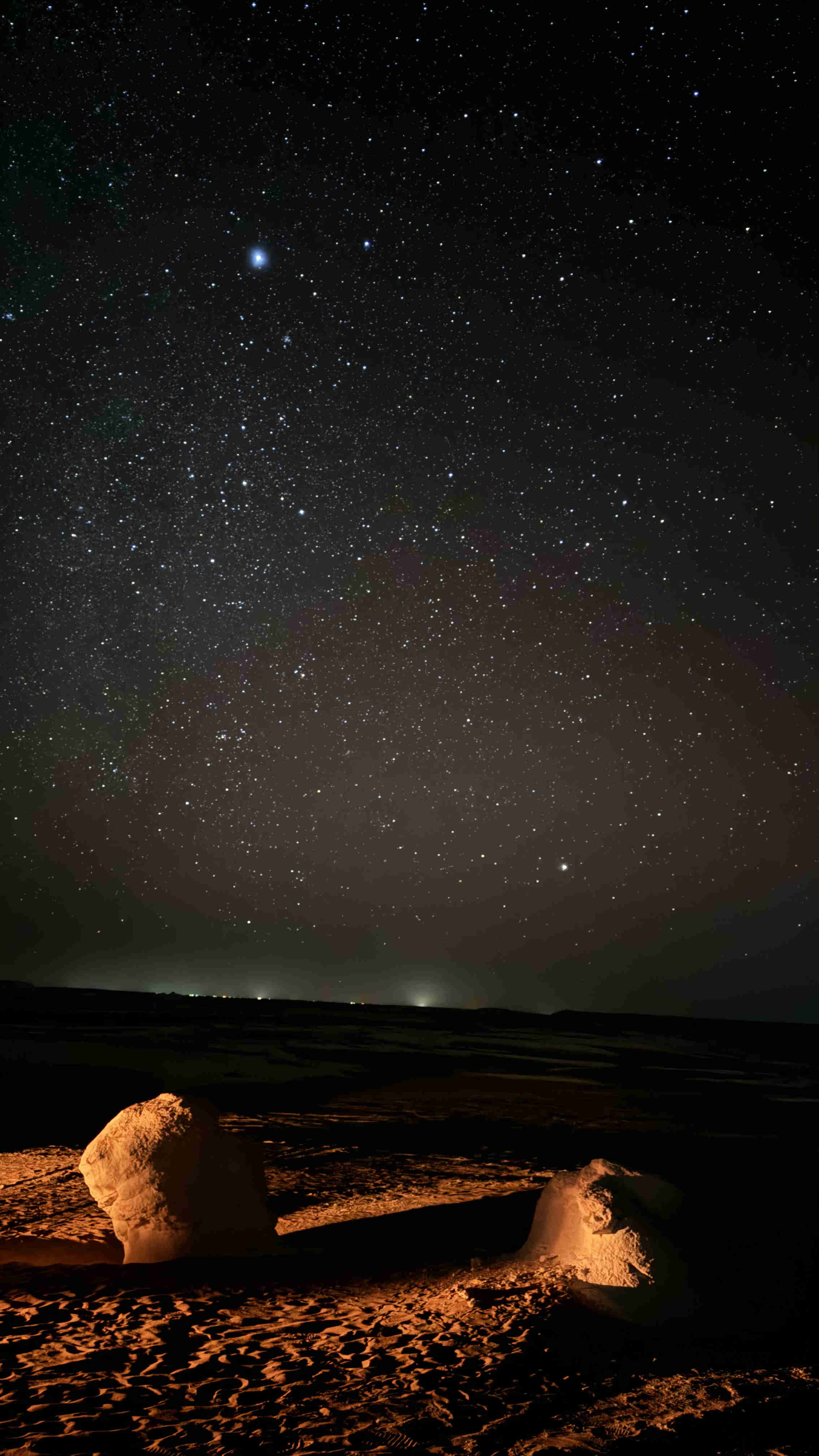 A vertical shot of the dark desert sky filled with bright stars and a wide stretch of the Milky Way, with two illuminated white rock formations in the foreground (White Desert night photography)