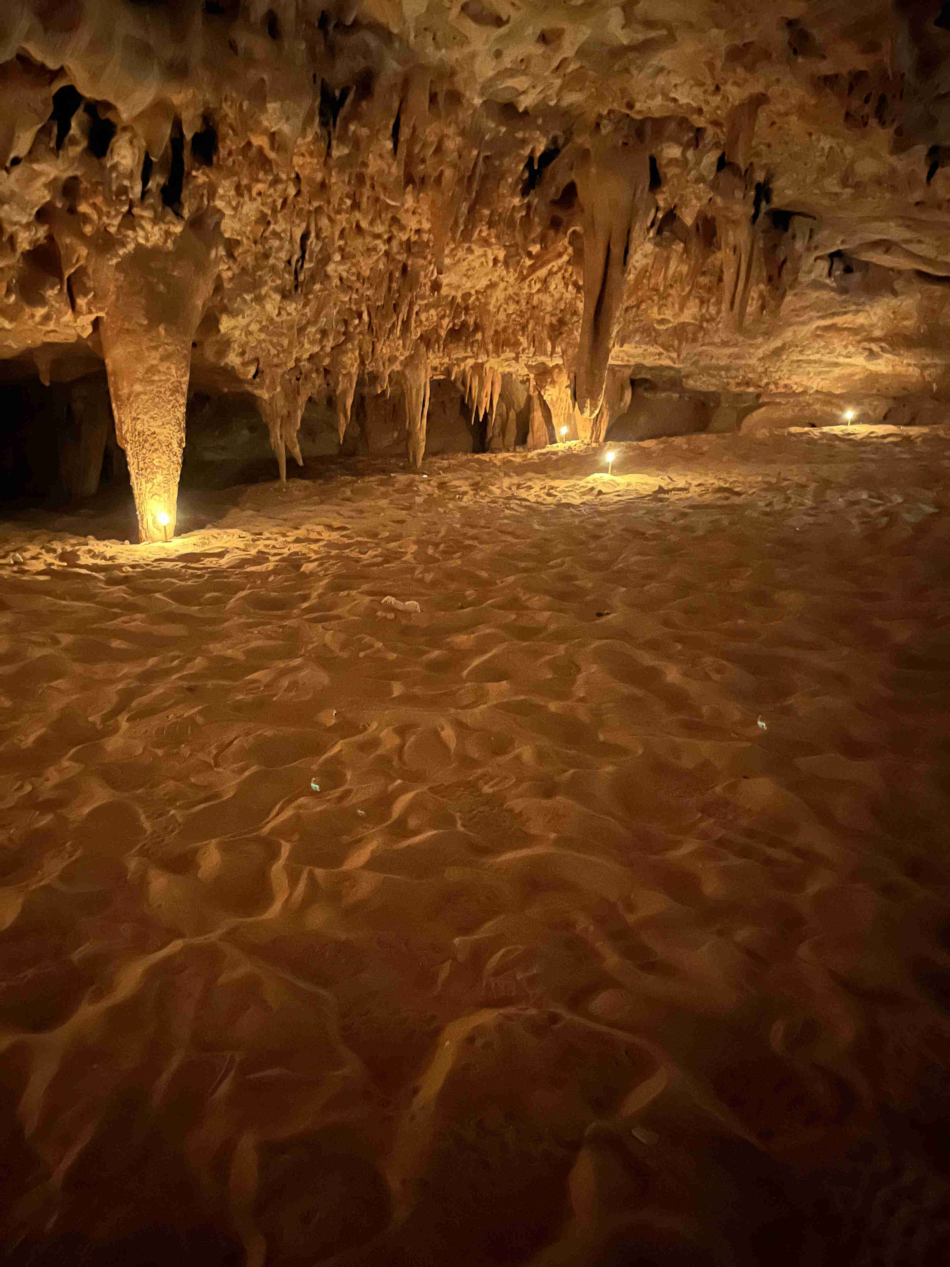 An interior view of Djara Cave lit by warm lamps, showing the sandy floor and dramatic stalactites hanging from the ceiling