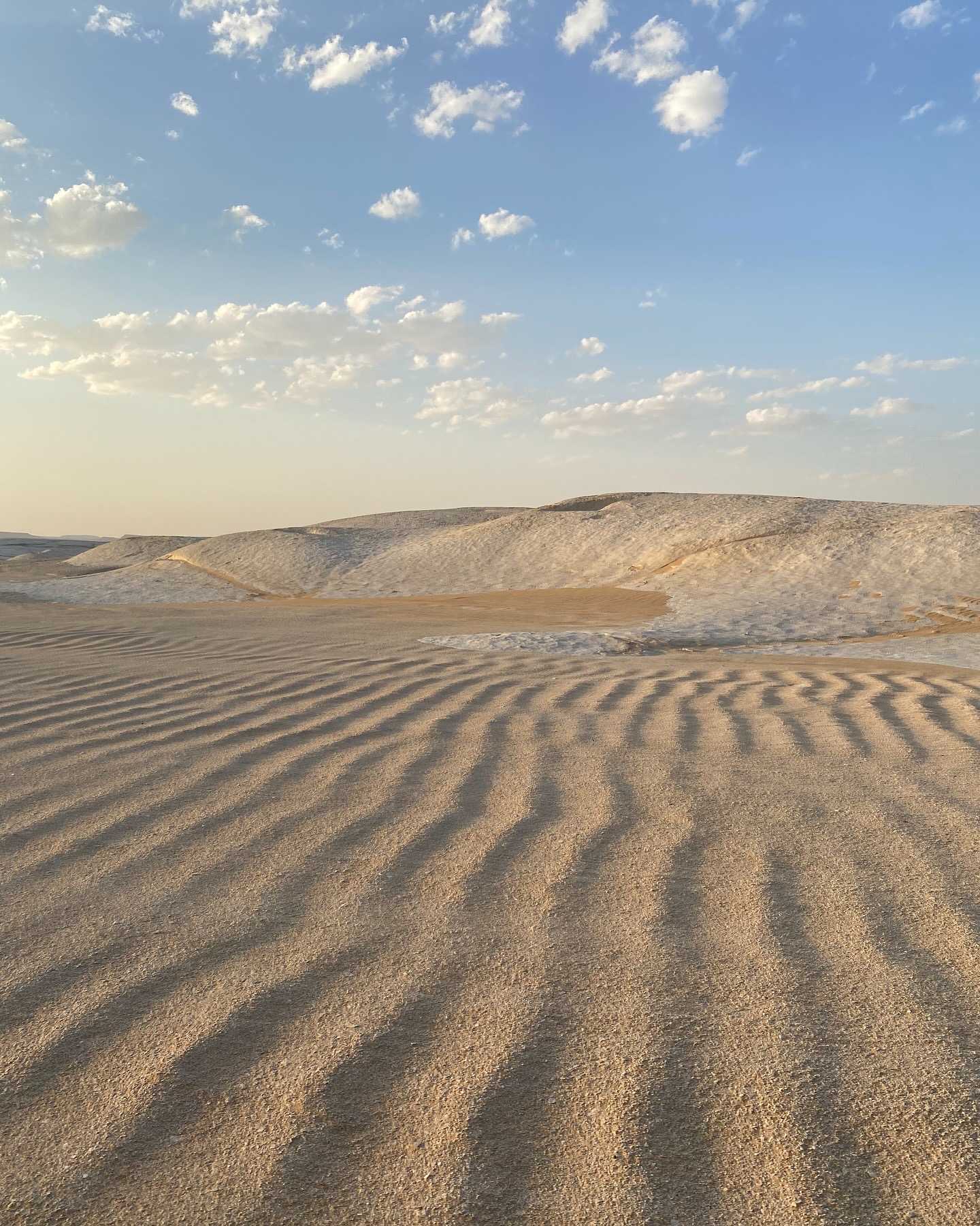 Wind-rippled sand dunes in the White Desert foreground leading up to white, chalk-covered hills under a blue sky with scattered clouds